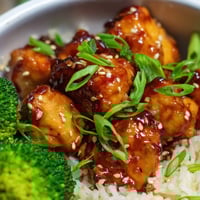 Fluffy steamed rice and tender broccoli florets support a bowl of sweet chili chicken, glistening with a tangy-sweet glaze and topped with fresh green onions.