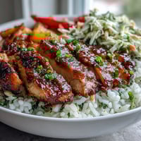 Glossy BBQ-glazed chicken slices rest on fluffy white rice next to crisp, creamy coleslaw and colorful roasted vegetables in a sturdy bowl.